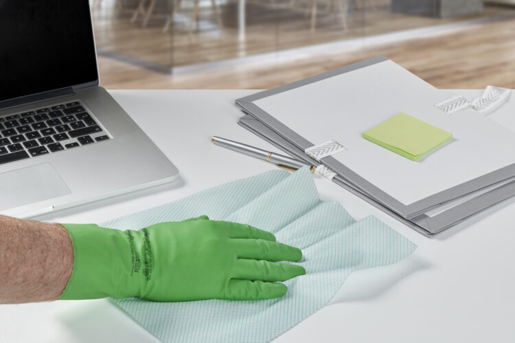 Una mano con un guante de goma verde pasa una bayeta blanca junto a un portátil plateado