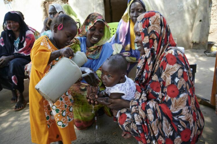 Una niña vierte agua sobre las manos de un bebé al que sujeta su madre.