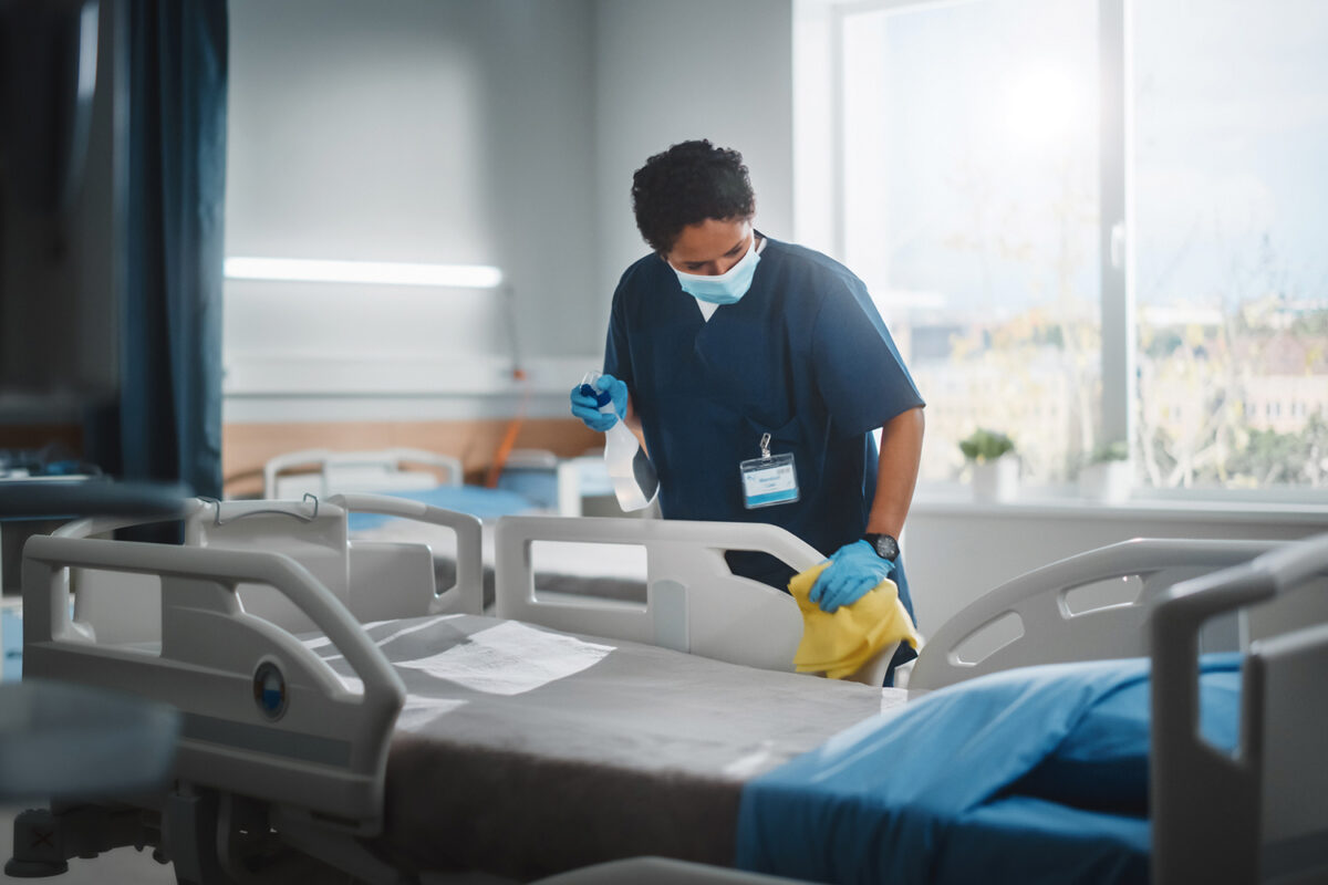 Hospital Ward: Professional Black Nurse Wearing Face Mask, Wiping the Bed, Cleaning Room After Covid-19 Patients Recover. Disinfection, Sterilizing, Sanitizing Clinic after Coronavirus Infected People