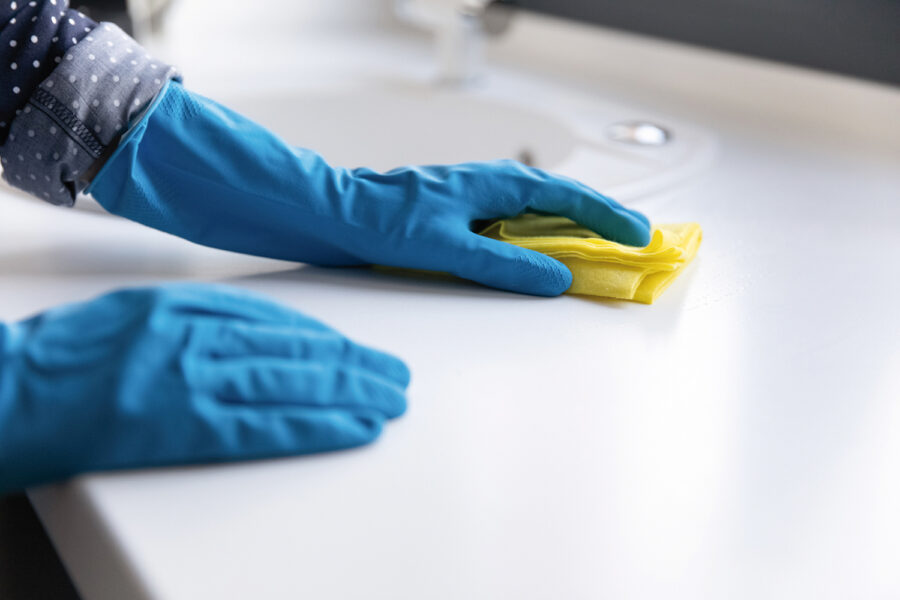 Housewife, cleaner, janitor wearing blue protective rubber gloves, washing table top or kitchen counter white surface with rag. Close up of hands. Household, cleaning service,